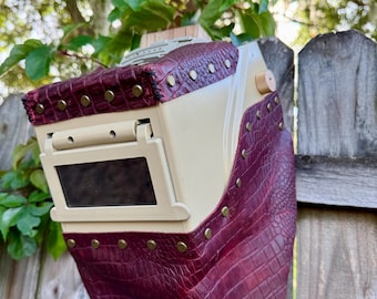 May include: A bespoke welding helmet, cream-coloured with burgundy crocodile-textured leather. It has a rectangular viewing window and brass-coloured rivets. The design is set against a wooden fence, with foliage in the background.