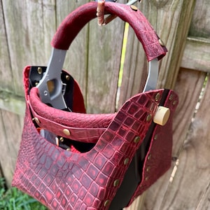 Op de afbeelding: Een lashelm met een rode hoes met een textuur van imitatie-alligatorhuid, messingkleurige klinknagels en een goudkleurige instelknop. De helm heeft een grijs intern frame en een gebogen handvat aan de bovenkant met zichtbare hardware.