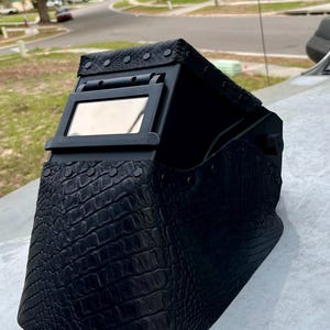 Pode incluir: Capacete de solda preto com um padr&atilde;o texturizado semelhante ao de um crocodilo. O capacete possui uma janela de visualiza&ccedil;&atilde;o retangular e &eacute; acentuado com rebites. Um equipamento de prote&ccedil;&atilde;o para soldagem.