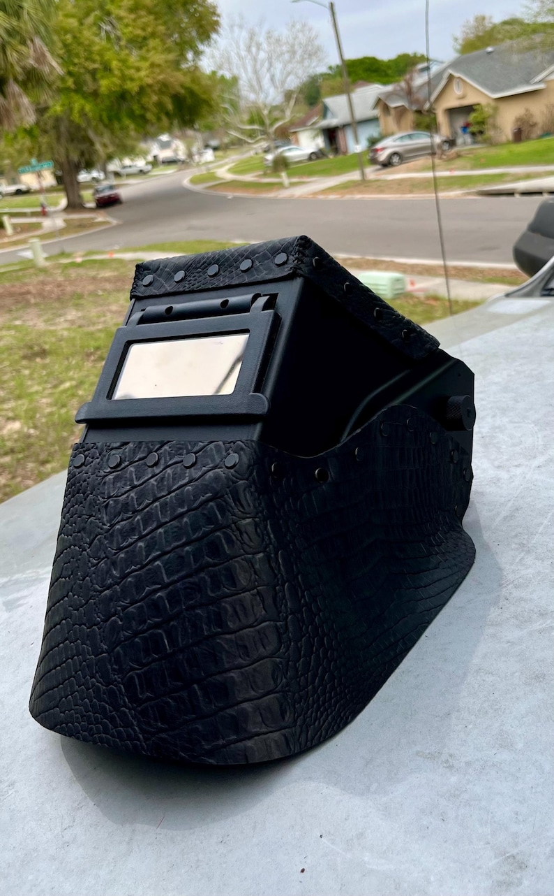 Pode incluir: Capacete de solda preto com um padr&atilde;o texturizado semelhante ao de um crocodilo. O capacete possui uma janela de visualiza&ccedil;&atilde;o retangular e &eacute; acentuado com rebites. Um equipamento de prote&ccedil;&atilde;o para soldagem.