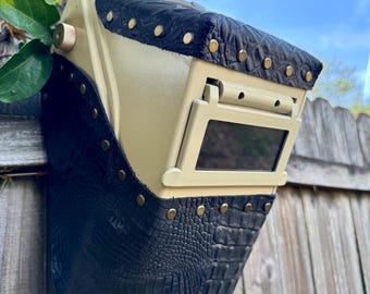 May include: A custom welding helmet with a beige body and black, textured leather detailing. The leather is secured with brass-coloured rivets. The helmet has a rectangular viewing window and is mounted on a wooden fence.