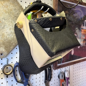 May include: A welding helmet featuring a cream-colored shell accented with black, textured, crocodile-patterned leather panels. The design includes brass-colored rivets and a circular adjustment knob on the side, providing a secure fit.