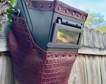 May include: A black welding helmet with a burgundy, crocodile-textured leather cover. The leather is secured with black rivets. The helmet has a rectangular viewing window and a control knob on the side. The background includes a wooden fence and greenery.