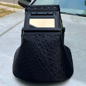Pode incluir: Um capacete de soldagem preto com um padr&atilde;o texturizado semelhante &agrave; pele de crocodilo. O capacete possui uma janela de visualiza&ccedil;&atilde;o retangular e foi projetado para proteger os olhos durante a soldagem. Rebites prendem o material texturizado &agrave; estrutura do capacete.