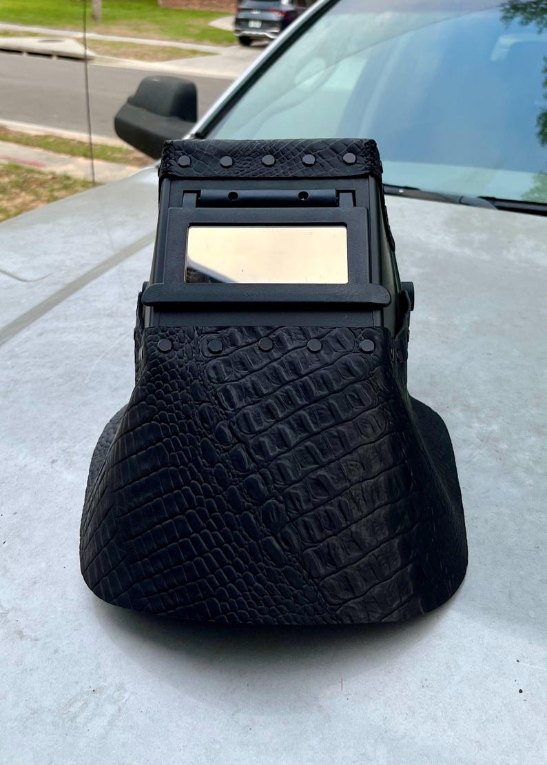 Pode incluir: Um capacete de soldagem preto com um padr&atilde;o texturizado semelhante &agrave; pele de crocodilo. O capacete possui uma janela de visualiza&ccedil;&atilde;o retangular e foi projetado para proteger os olhos durante a soldagem. Rebites prendem o material texturizado &agrave; estrutura do capacete.