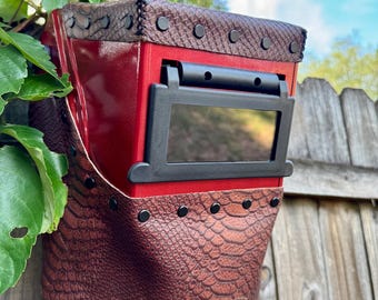 May include: A red welding helmet with a brown, snakeskin-textured leather face shield and top. The helmet has a black viewing window and is secured with black rivets. The background includes green foliage and a wooden fence.