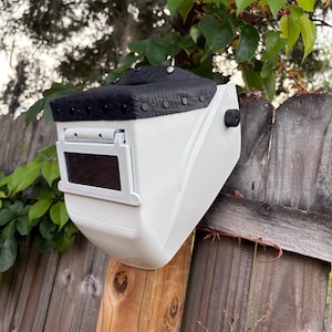 May include: A white welding helmet with a black padded top and a rectangular viewing window. The helmet is mounted on a wooden fence post, with green leaves and a wooden fence in the background.