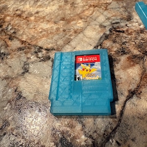 Puede incluir: Una funda de cartucho de juego de Nintendo Switch azul claro impresa en 3D. La funda contiene un cartucho de juego con un logotipo rojo de Nintendo Switch y un gráfico de Pikachu. El juego está clasificado "E" para todos.