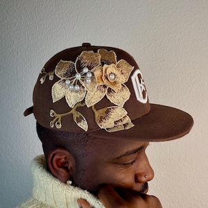 Op de afbeelding: Een bruine baseballcap met geborduurde goud- en bruine bloemen en imitatieparels. De cap heeft een gebogen klep en een wit logo aan de zijkant. Het bloemendesign bevindt zich aan de voorkant.