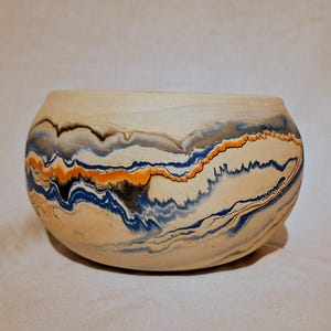 May include: A rounded ceramic bowl featuring a marbled pattern with swirling bands of cream, deep blue, bright orange, and charcoal gray. The surface has a matte finish with organic, flowing lines that wrap horizontally around the entire vessel.
