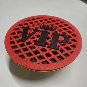 May include: A red, circular object with a lattice design. The word "VIP" is displayed in black, textured letters, topped with a crown. The object has a gold-coloured rim.