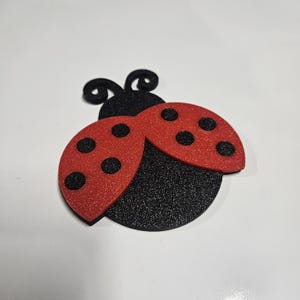 May include: A glittery ladybug decoration with a red body and black spots and a black body. The ladybug has two antennae. The ladybug is a craft item.