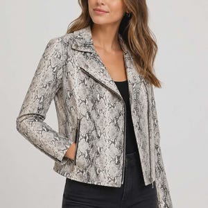May include: A grey and white faux snakeskin jacket. The jacket has a classic biker design with a notched collar, an asymmetrical zip, and zipped pockets. The model is wearing a black top and black jeans.