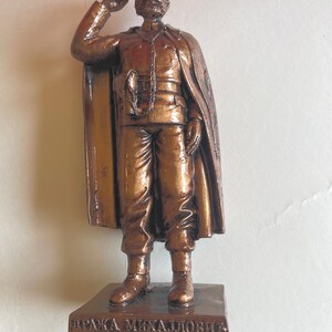 May include: A bronze-colored statue of a man in a military uniform, saluting. The figure has a beard and is wearing a hat and cape. The statue is mounted on a rectangular base with engraved text.
