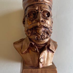 May include: A bronze-colored bust of a man with a mustache, beard, and glasses. He is wearing a hat and a uniform with a buttoned collar. The bust is mounted on a rectangular base.