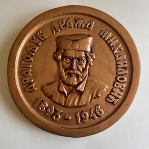 May include: A bronze-colored circular medallion featuring a portrait of a man with a beard, glasses, and a hat. The text "ДРАЖА МИХАИЛОВИЋ" and the years "1893-1946" are inscribed around the portrait. The medallion has a raised relief design.