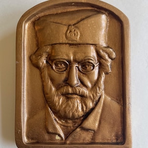 May include: A bronze-colored relief sculpture of a man with glasses, a beard, and a hat. The rectangular plaque has a rounded top and a detailed depiction of the man's face and attire. The sculpture is likely a portrait or commemorative piece.