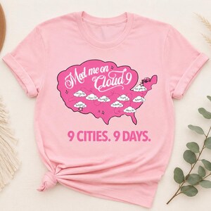 May include: A light pink t-shirt featuring a pink map graphic with the text "Meet me on Cloud 9" and "9 Cities. 9 Days." The map includes cloud illustrations and city names. The shirt has short sleeves and a crew neck.