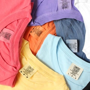 May include: A close-up shot of a collection of Comfort Colors t-shirts in various colors, including coral, purple, orange, yellow, and blue. Each shirt has a tag with the brand name and size information.