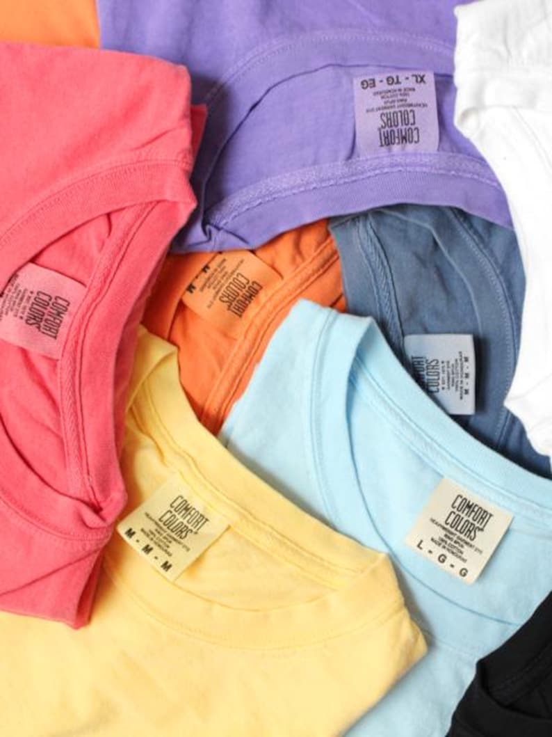 May include: A close-up shot of a collection of Comfort Colors t-shirts in various colors, including coral, purple, orange, yellow, and blue. Each shirt has a tag with the brand name and size information.