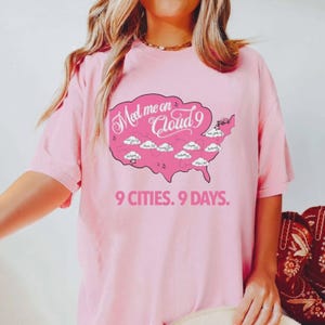 May include: Pink t-shirt with a graphic of the United States map in a cloud shape, featuring the text "Meet me on Cloud 9" and "9 Cities. 9 Days." The map includes city names. The shirt is a light pink color.