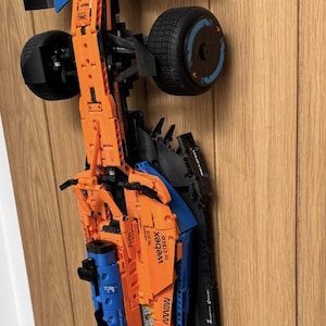 Könnte beinhalten: Ein orange-blaues Modellrennauto mit schwarzen Reifen, montiert auf Holz. Das Modell hat Sponsorenlogos und den Text "McLaren" und "webex by cisco". Detaillierte Nachbildung eines Rennwagens.