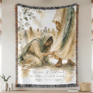 May include: A woven throw blanket featuring a watercolor-style illustration of a person kneeling to touch the hem of a robe. The text reads, "She said, 'If I just touch his clothes, I will be healed.' MARK 5:28." The blanket has a fringed edge.