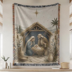 May include: A woven tapestry blanket featuring a nativity scene with a central stable, a star above, and figures of a person in a blue robe and a person in a brown robe kneeling beside a manger with a baby, framed by a fringed edge.