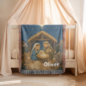 Könnte beinhalten: Eine gewebte Wohndecke mit einer Krippenszene, einem Stern, einem Stall und den Figuren von Maria, Josef und dem Jesuskind in einer Krippe. Das Design zeigt ein Lamm, einen Esel und eine Kuh, mit dem Namen Oliver in weißer Schrift am unteren Rand.