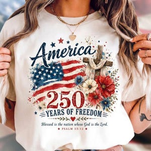 Puede incluir: Una camiseta color crema con un gráfico que incluye el texto America 250 Years of Freedom y Psalm 33:12. El diseño presenta una bandera estadounidense en acuarela, una cruz de madera con tela blanca y flores rojas, blancas y azules.