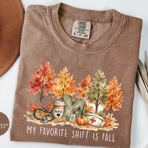 May include: An espresso-colored t-shirt featuring a graphic of autumn trees, a stethoscope, a coffee cup labeled Nurse, a scrub top, a pumpkin, and a nurse cap. The text reads MY FAVORITE SHIFT IS FALL. The shirt has a Comfort Colors brand neck label.