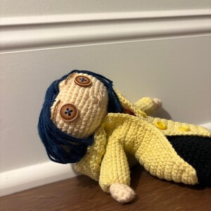 Puede incluir: Mu&ntilde;eca de ganchillo hecha a mano con una cara beige claro, ojos de bot&oacute;n y cabello azul oscuro. La mu&ntilde;eca lleva un abrigo amarillo y pantalones negros. La mu&ntilde;eca est&aacute; tumbada sobre una superficie de madera.