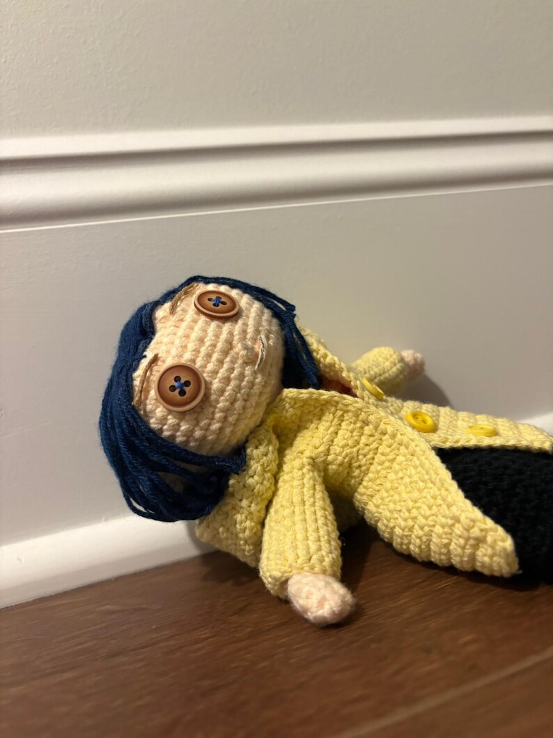 Puede incluir: Mu&ntilde;eca de ganchillo hecha a mano con una cara beige claro, ojos de bot&oacute;n y cabello azul oscuro. La mu&ntilde;eca lleva un abrigo amarillo y pantalones negros. La mu&ntilde;eca est&aacute; tumbada sobre una superficie de madera.