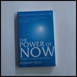 May include: A blue book titled The Power of Now: A Guide to Spiritual Enlightenment by Eckhart Tolle. The cover features a bright white light radiating from the top center against a blue background, with text stating it is an international bestseller.