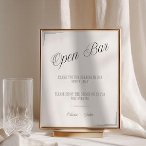 May include: A rectangular sign in a gold frame with the text Open Bar, Thank you for sharing in our special day, Please enjoy the drinks on us for this evening, and the names Olivia and John, accompanied by two clear textured glass tumblers.