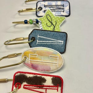 May include: A collection of personalized keychains with embroidered letters. Each keychain features a unique design, including denim, floral, and cow print patterns. The keychains have gold-colored clasps and are arranged in a vertical row.