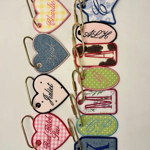 May include: A collection of embroidered fabric keychains with gold-toned metal rings. The shapes include hearts, stars, ovals, and rectangles, featuring various patterns like gingham, denim, cow print, and floral, with personalized names and letters.