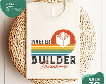 May include: A cream-colored t-shirt featuring a graphic with a white building block, a red sun, and horizontal stripes in yellow, green, and blue. The text reads MASTER BUILDER Theodore in a distressed font style with a retro aesthetic.