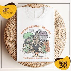 A white t-shirt with a graphic design depicting various Disney landmarks and elements, including Mickey Mouse and the Eiffel Tower, along with the text "Walt Disney World" and "The Happiest place in earth".
