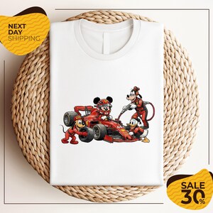 May include: A white t-shirt featuring a graphic print of cartoon characters in red racing gear servicing a red Formula One race car. Mickey Mouse sits in the cockpit, while Goofy and Donald Duck assist with the tires and fuel hose.