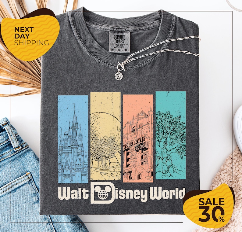 A gray t-shirt with a graphic design featuring various Disney landmarks, including the Eiffel Tower, the Walt Disney World resort, and a tree. The t-shirt is displayed on a white background, and there are jeans and a sweater visible in the background.