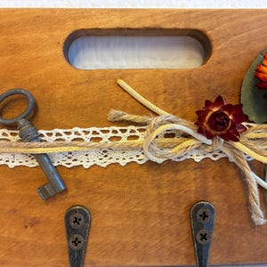 Könnte beinhalten: Schlüsselhalter aus Holz mit Griff, mit einem antiken Schlüssel, Spitze und Schnurakzenten. Verziert mit einem Trockenblumenarrangement in Orange- und Rottönen. Enthält Metallhaken für Schlüssel.