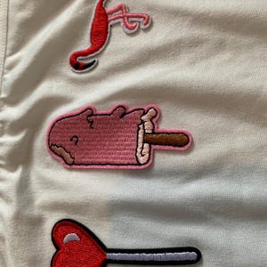 以下が含まれることがあります： 白い生地に刺繍されたパッチ。パッチには、赤いフラミンゴ、ピンクのアイスキャンディー、赤いハート型のロリポップが含まれています。パッチには白と黒の縁取りがあり、衣類やアクセサリーに適しています。