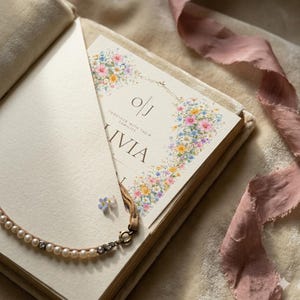 May include: A wedding invitation with floral accents and the names "O/J" and "Olivia" printed on the front. The invitation is nestled in a cream-colored book, with a pearl necklace and a pink ribbon nearby.