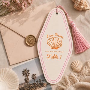 Puede incluir: Un letrero de mesa en forma de diamante de color blanco roto con bordes rosas y una borla rosa. El letrero presenta una ilustración naranja de una concha, el texto "Lane Moore" y "Table 1" en naranja, y un agujero superior para la borla.