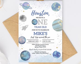 Pode incluir: Um convite de aniversário com tema espacial. O convite apresenta planetas, estrelas e uma ilustração de foguete. O texto diz "Houston, we have a one year old" e detalhes da festa. Em um fundo branco com um envelope marrom.