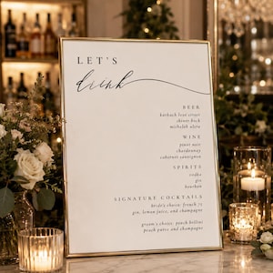 May include: A rectangular gold-framed sign with a white background featuring the text LET'S drink in black lettering. The menu lists beer, wine, spirits, and signature cocktails including bride's choice french 75 and groom's choice peach bellini.