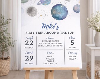Pode incluir: Um letreiro personalizado em um cavalete com tema espacial, com planetas, um foguete e o texto "Mike's First Trip Around The Sun". Ele lista o peso da criança como 10 kg e a altura como 74 cm.