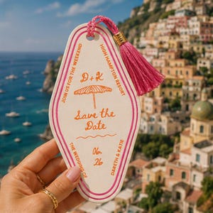 May include: A cream-colored, retro-style hotel key tag with pink trim and a pink tassel. The tag reads "Save the Date" with the names Dareth & Katie and the date 06.26. The background shows a coastal town.