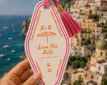 Pode incluir: Uma etiqueta de chave de hotel em estilo retrô creme com acabamento rosa e uma borla rosa. A etiqueta diz "Save the Date" com os nomes Dareth & Katie e a data 06.26. O fundo mostra uma cidade costeira.